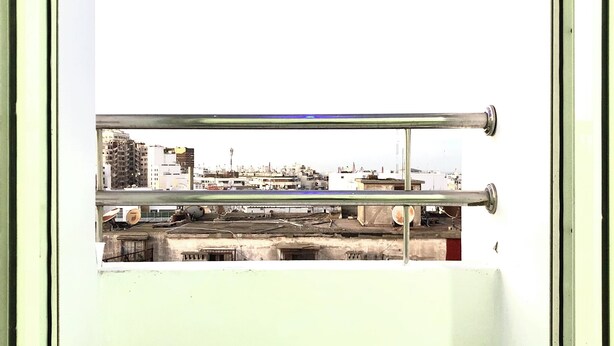A small balcony with a metal railing, a stool, and a view of a cityscape through a window. - Chinese guesthouses in Casablanca (Casablanca)