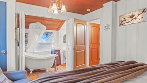A bedroom with a large bed, a wooden ceiling, a white bathtub, and a door leading to another room.