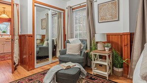 A cozy living room with a grey armchair, a small ottoman, a wooden side table with a lamp, and a potted plant.