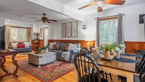 A spacious living room with a sofa, coffee table, and dining area. There are ceiling fans, a large window with blinds, and a decorative wall piece.