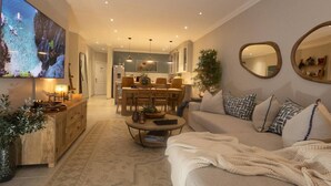 A modern living room with a large flat-screen TV, a dining area, and a cozy seating area with a sofa and cushions.