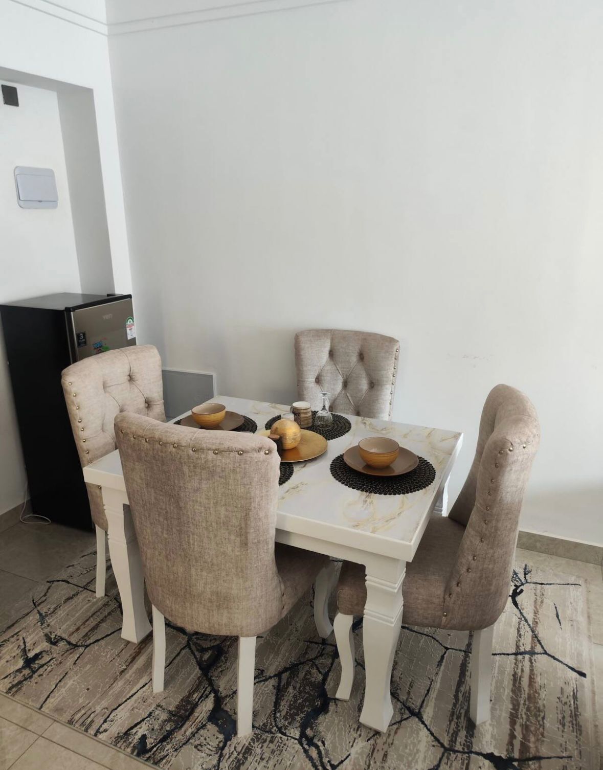 Uma área de jantar com uma mesa branca, quatro cadeiras estofadas e uma geladeira preta.