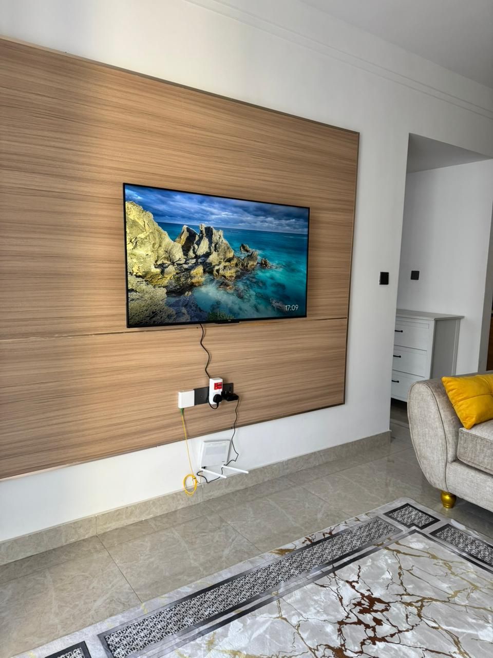 Ein modernes Wohnzimmer mit einem an der Wand montierten Fernseher, der ein Küstenpanorama zeigt, einer Holzwand, einem grauen Sofa mit einem gelben Kissen und einem gemusterten Teppich.