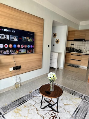 A modern kitchen with wooden cabinets, a built-in TV displaying streaming apps, a white dining area with a round table, and a decorative vase with flowers.