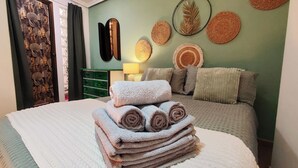 Ein ordentlich bezogenes Bett mit aufgerollten Handtüchern, eine grüne Wand mit Wandweberei und ein Spiegel.