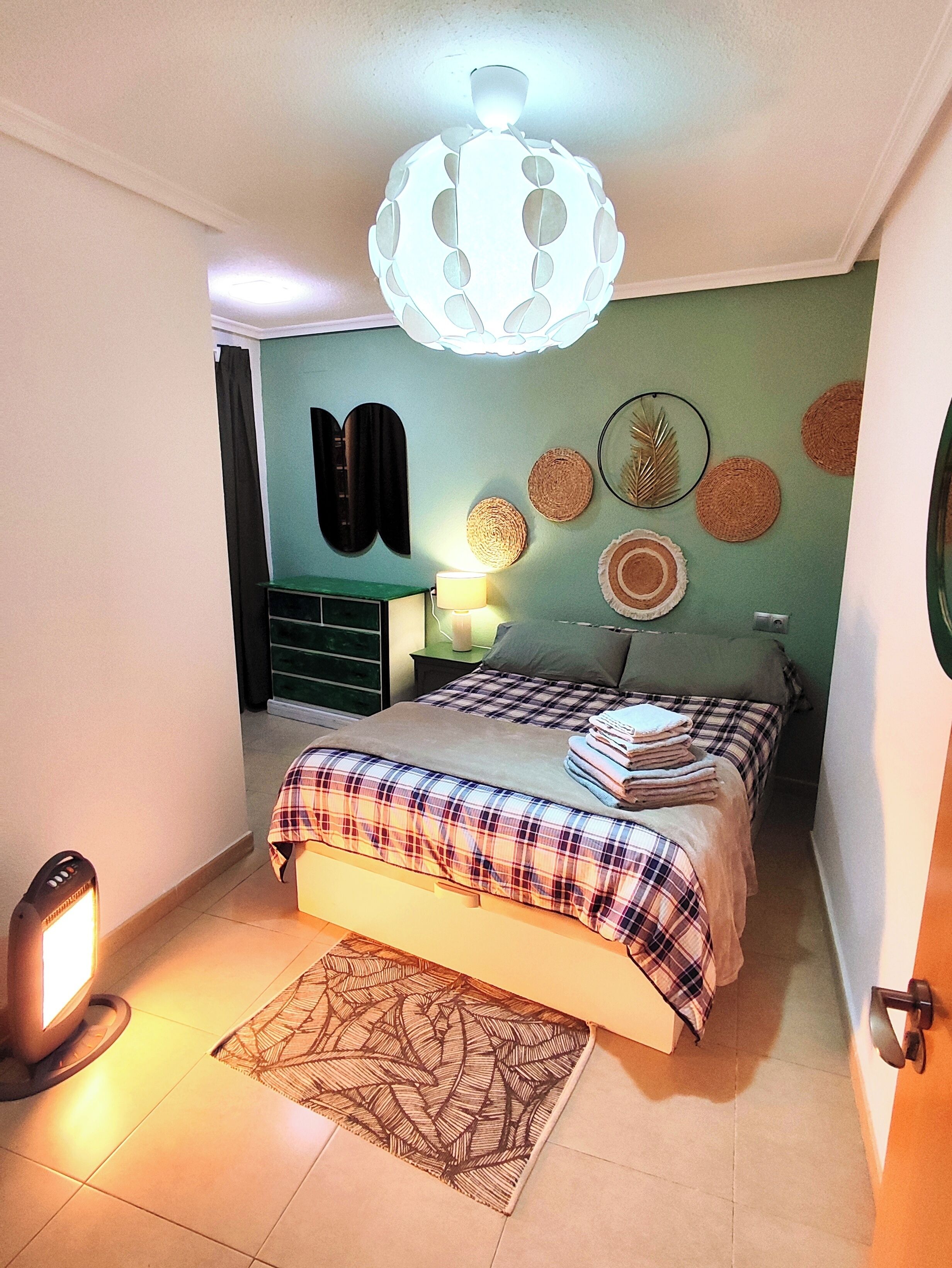 A bedroom with a bed, a nightstand, a lamp, a wall-mounted mirror, and a ceiling light fixture.