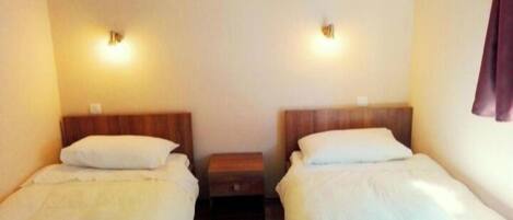 A room with two single beds, each with a wooden headboard and white bedding. There is a small wooden nightstand between the beds.