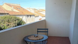 Uma pequena varanda com uma mesa e duas cadeiras, vista de prédios ao fundo e piso de azulejos.