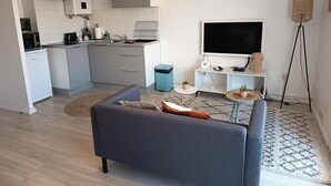 Cozinha moderna com armários brancos, micro-ondas e pia. Há um sofá cinza com almofadas e uma pequena mesa com uma planta. Uma televisão está fixada na parede, e um abajur é visível.