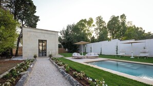 Un espace piscine extérieur moderne, avec un chemin de pierre menant à la piscine, entouré d’une pelouse bien entretenue et d’un bâtiment blanc doté d’une porte en verre.