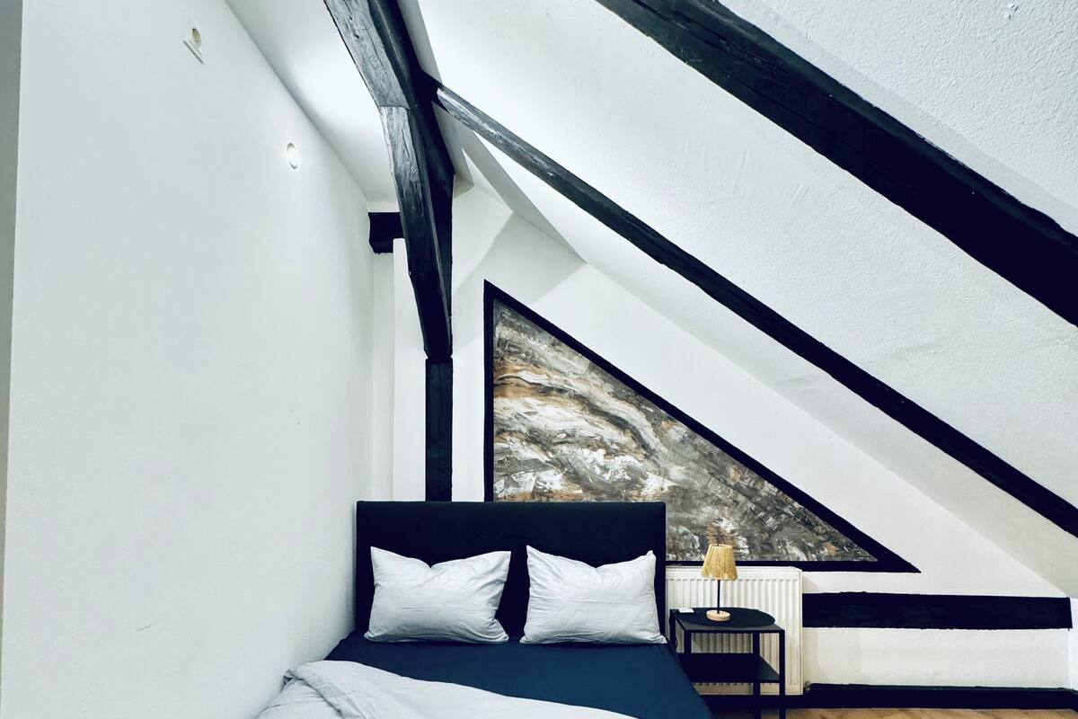 A bedroom with a sloped ceiling, a bed with white bedding, a nightstand, and a lamp.