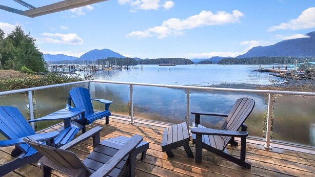 Een terras met houten stoelen met uitzicht op een rustig meer met bootjes en steigers, omgeven door bergen en begroeiing.