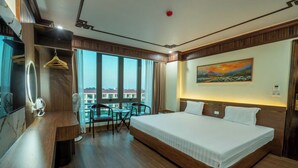 Ein Hotelzimmer mit einem großen Bett, einem Schreibtisch mit Stuhl, einem kleinen Tisch mit Stühlen und einem großen Fenster mit Blick auf Gebäude.
