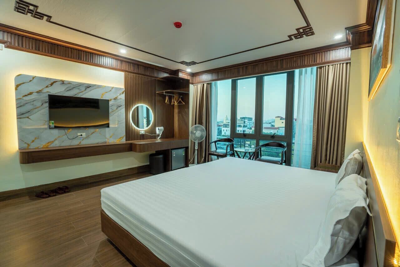 A hotel room with a large bed, a TV mounted on a marble wall, a small desk with a mirror, and a view of the city through large windows.