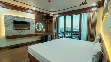 A hotel room with a large bed, a TV mounted on a marble wall, a small desk with a mirror, and a view of the city through large windows.