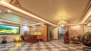 Eine Hotelhalle mit Schachbrettmuster-Boden, einer Rezeption, einem großen Fernsehbildschirm und traditioneller Einrichtung.