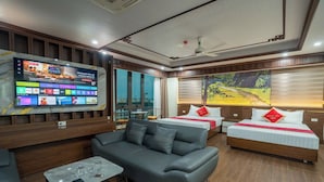 Ein Hotelzimmer mit einer Couch, zwei Betten, einem Fernseher, der Streaming-Dienste anzeigt, und einem großen Fenster mit einem Gemälde.