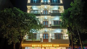Ein mehrstöckiges Hotel mit beleuchteter Beschilderung, einem gut beleuchteten Eingangsbereich und einer angelegten Grünfläche mit Bäumen.