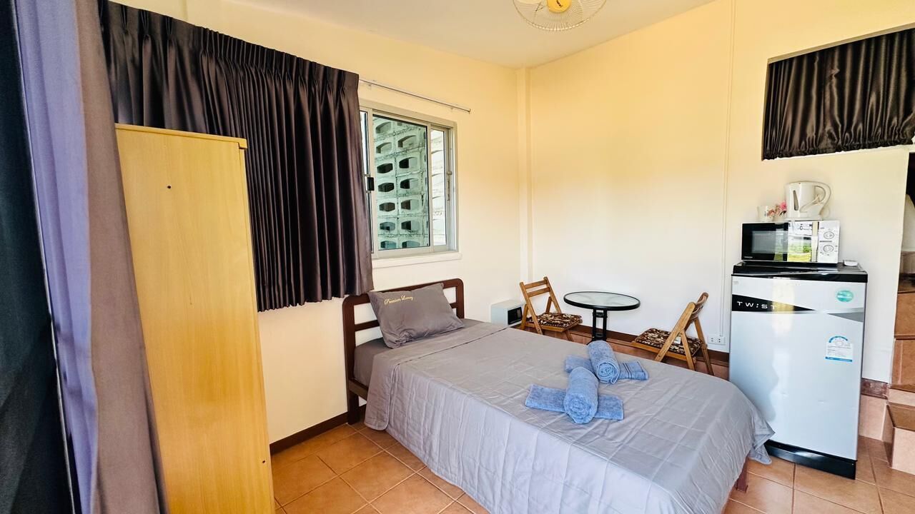 A bedroom with a bed, a small table, a chair, a wardrobe, a window with blinds, and a kitchenette with a microwave and refrigerator.