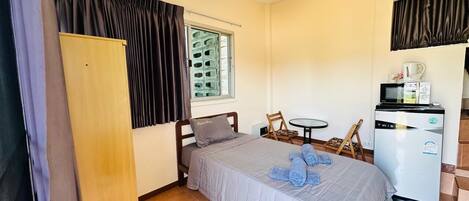 A bedroom with a bed, a small table, a chair, a wardrobe, a window with blinds, and a kitchenette with a microwave and refrigerator.