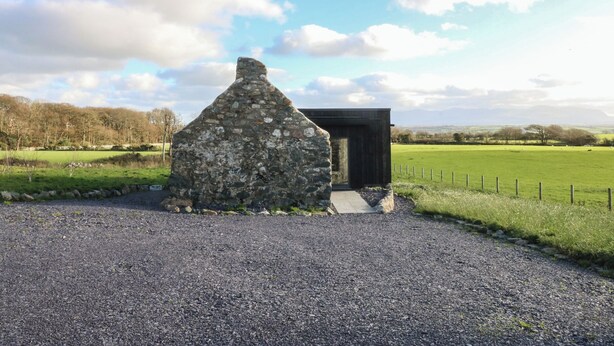A stone structure with a wooden extension, surrounded by a gravel path and a fenced field.