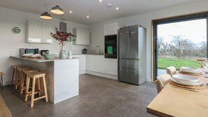A modern kitchen with white cabinets, a central island with stools, a stainless steel refrigerator, and a view of the outdoors through a large window. - Tithe Barn Lodge (Bodorgan)
