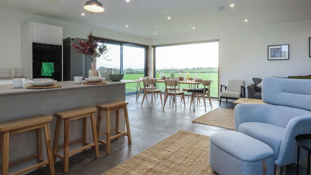 A modern kitchen with a large island, wooden stools, and a dining area with a view of the countryside. - Tithe Barn Lodge (Bodorgan)