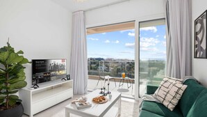 Un salon moderne avec une grande télévision, un canapé vert, une table basse sur laquelle sont disposées des pâtisseries, et un balcon offrant une vue sur la ville.