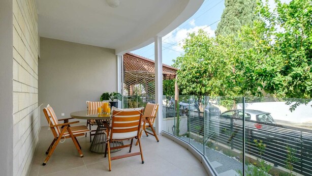 A balcony with a table and chairs, a potted plant, and a view of a tree and street. - Casa Centrale by Nomads (Paphos)