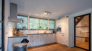 A modern kitchen with wooden cabinets, a built-in oven, and a large window with plants. - Lovely villa on the edge of the forest (Den Burg)