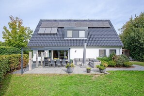 A house with a sloped roof, solar panels, a covered patio with outdoor furniture, and a well-maintained garden. - Lovely villa on the edge of the forest (Den Burg)