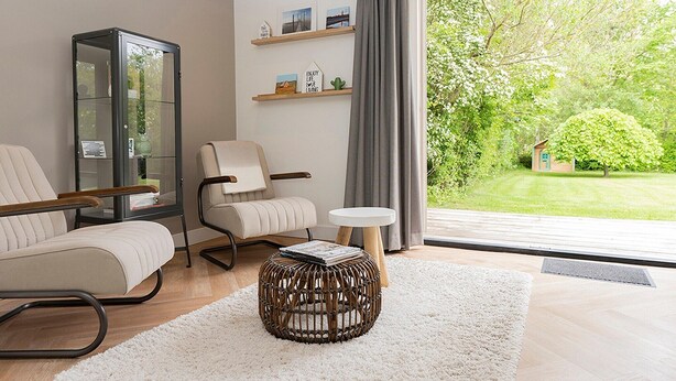 Ein modernes Wohnzimmer mit einer Glasschrank, zwei Sofas, einem runden Rattan-Hocker und Blick auf einen gepflegten Garten durch große Fenster.