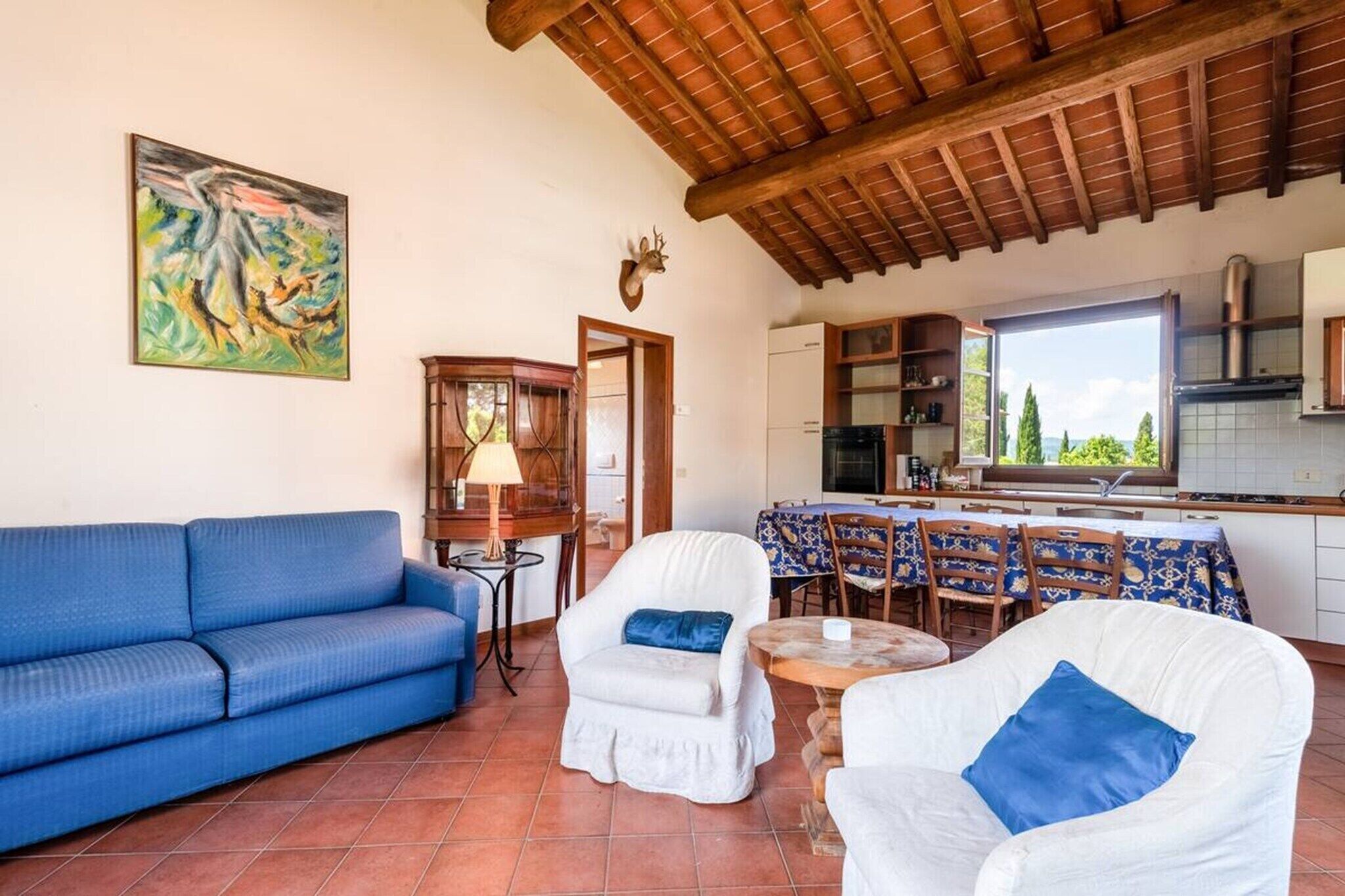 A living room with a blue sofa, white armchairs, a dining table with chairs, a kitchen area, and a mounted deer head.