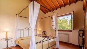 Ein Bett mit Baldachin, weiße Vorhänge, Holzbalkendecke, ein Fenster mit Blick auf Bäume und zwei Nachttische mit Lampen.