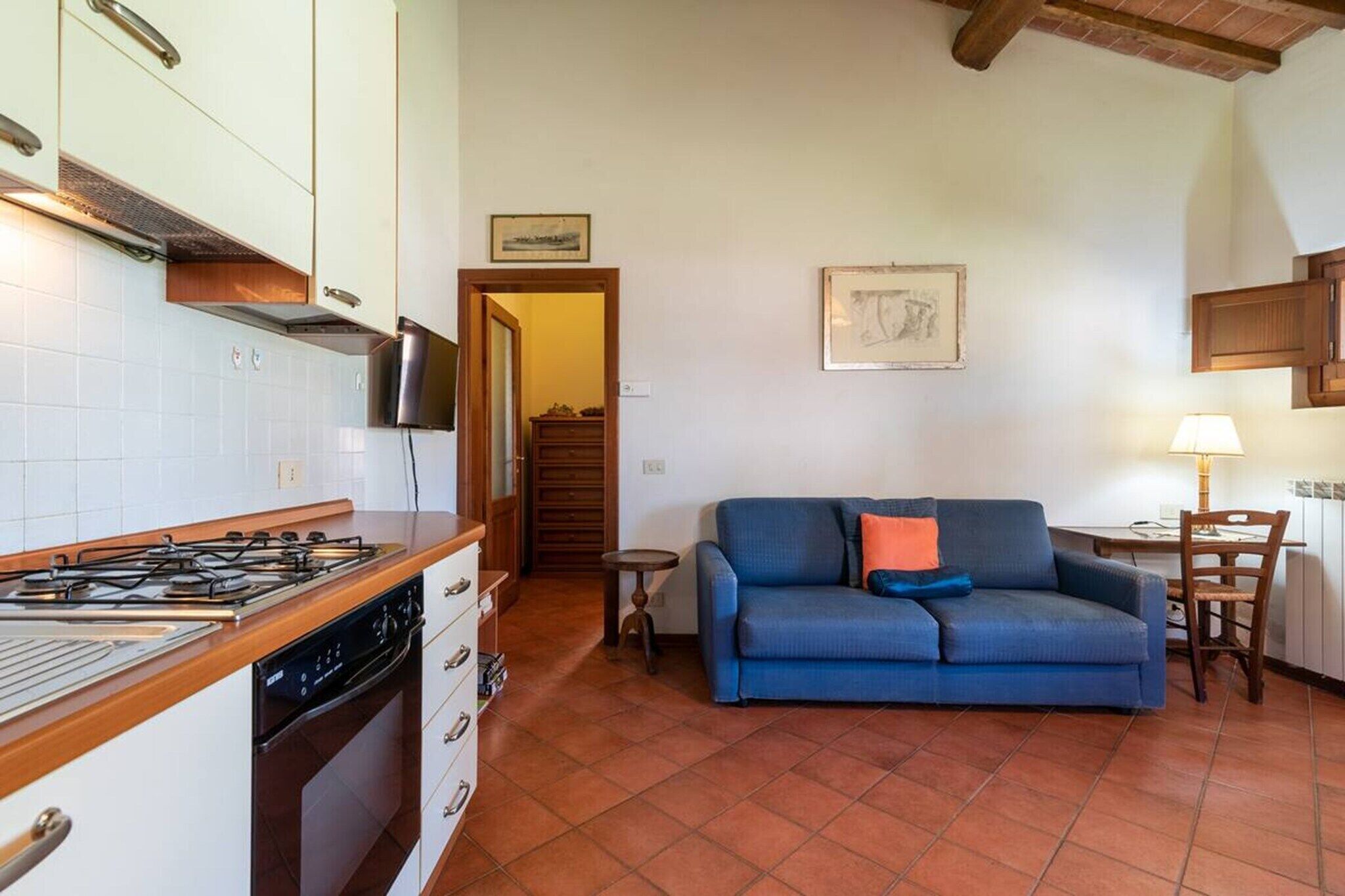 A compact kitchen with a gas stove, a dishwasher, and white cabinets. There is a blue sofa with a cushion and a small table with a lamp.