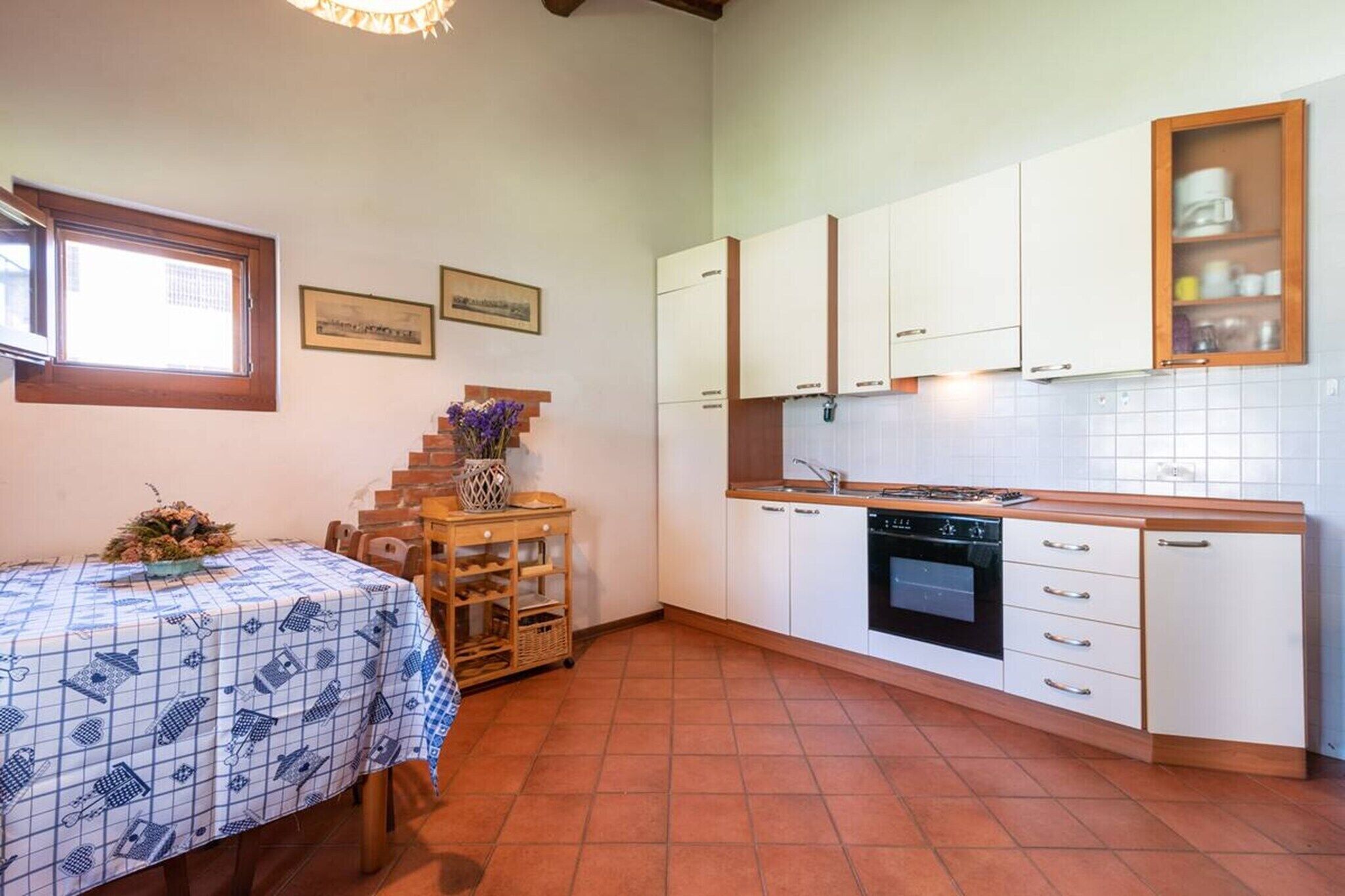 A compact kitchen with white cabinets, a stove, and a tiled backsplash. There is a dining table with a patterned tablecloth and a small wooden side table with a vase.