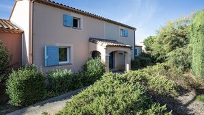 A two-story house with a tiled roof, a front entrance with a blue door, and a small porch area. - P. Minervois J N°03 - Villa 3P6 (Homps)