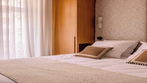 Un lit bien fait, avec du linge de lit blanc et des coussins à motifs, une tête de lit en bois et une grande fenêtre ornée de rideaux légers.