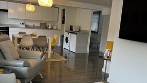 Een moderne woonkamer met een grijze bank, een salontafel en een vloerkleed met een patroon. Er is een keukenhoek met een wasmachine en een eethoek met stoelen.