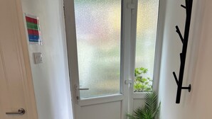 Een gang met een houten vloer, een witte deur, een raam met uitzicht op groen, een potplant en een zwarte kapstok.