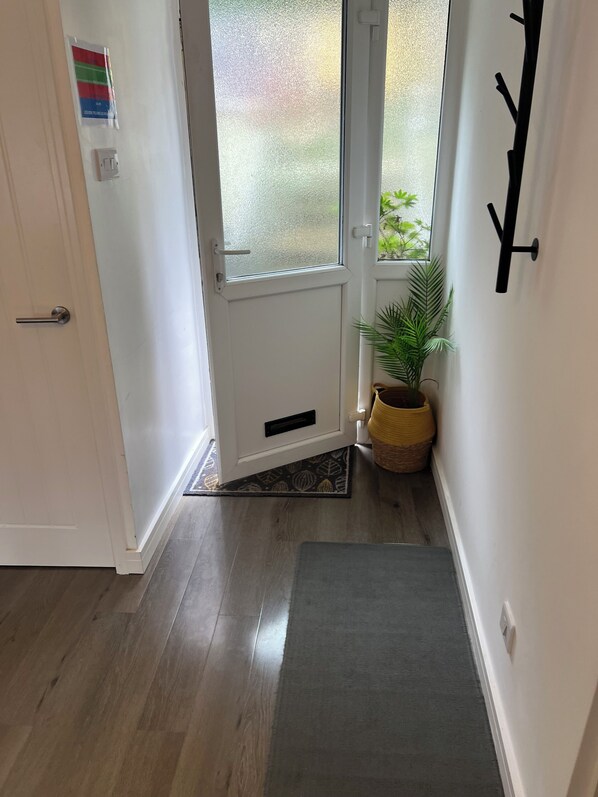 Un couloir avec un plancher en bois, une porte blanche, une fenêtre donnant sur la verdure, une plante en pot et un porte-manteau noir.