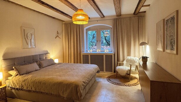 A bedroom with a large bed, wooden beams, a hanging light fixture, a window with curtains, and a bedside table with a lamp. - Family- & dog-friendly apartment near the Elbe with direct access to the park (Dresden)
