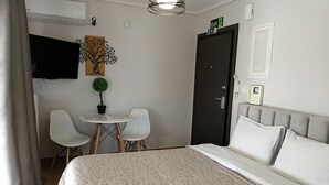 Quarto de hotel com uma cama, uma mesa redonda pequena com cadeiras, um vaso de planta, uma televisão na parede e um sinal de saída.