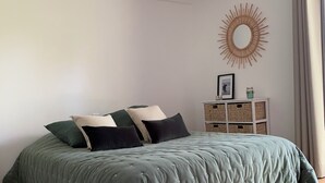 Quarto com cama, piso de madeira, parede branca, espelho de sol decorativo e um pequeno armário branco.