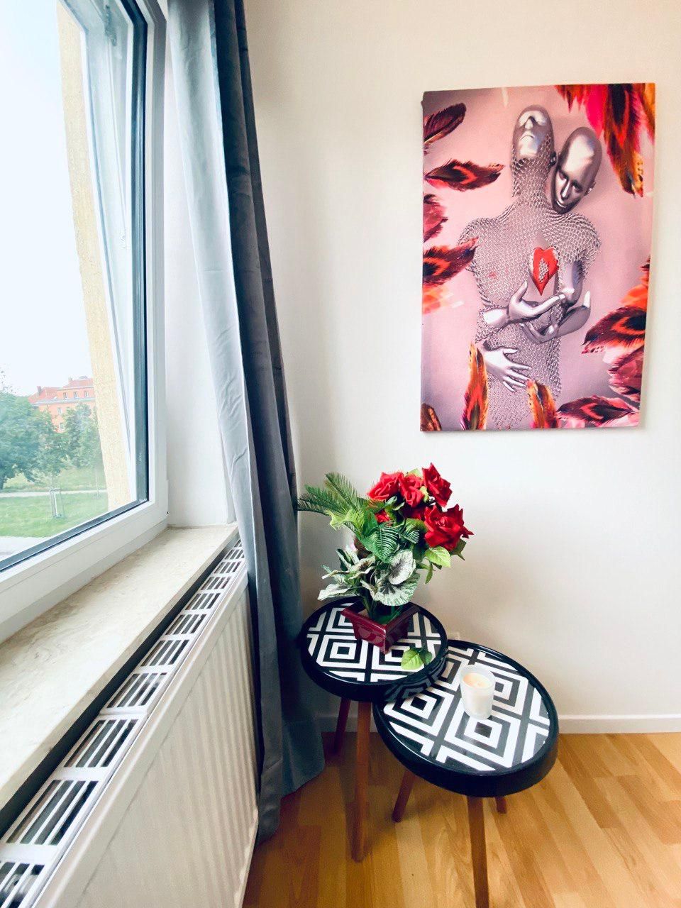 A room with a radiator, a window with curtains, a wall-mounted artwork, and a small table with a vase of flowers.