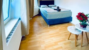 A bedroom with a bed, a window with curtains, a radiator, a wooden floor, and a small table with a vase of flowers. - Budget Friendly 4 BR Apt for Groups (Vienna)