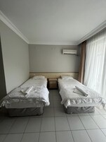 Una habitación de hotel con dos camas individuales, una mesita de noche de madera, un equipo de aire acondicionado y un ventanal con cortinas traslúcidas.