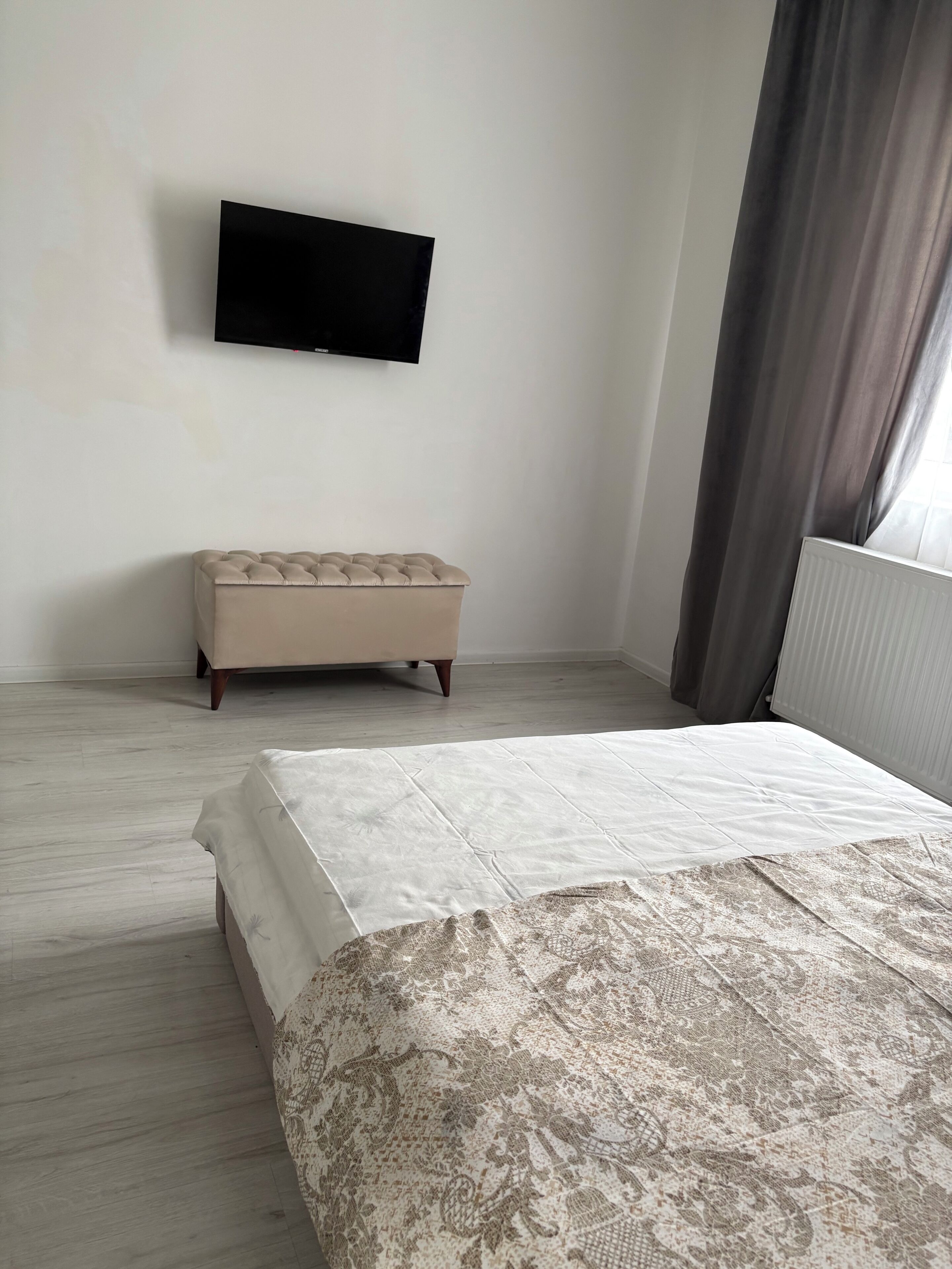 A bedroom with a bed, a tufted ottoman, a flat-screen TV on the wall, and a window with curtains.