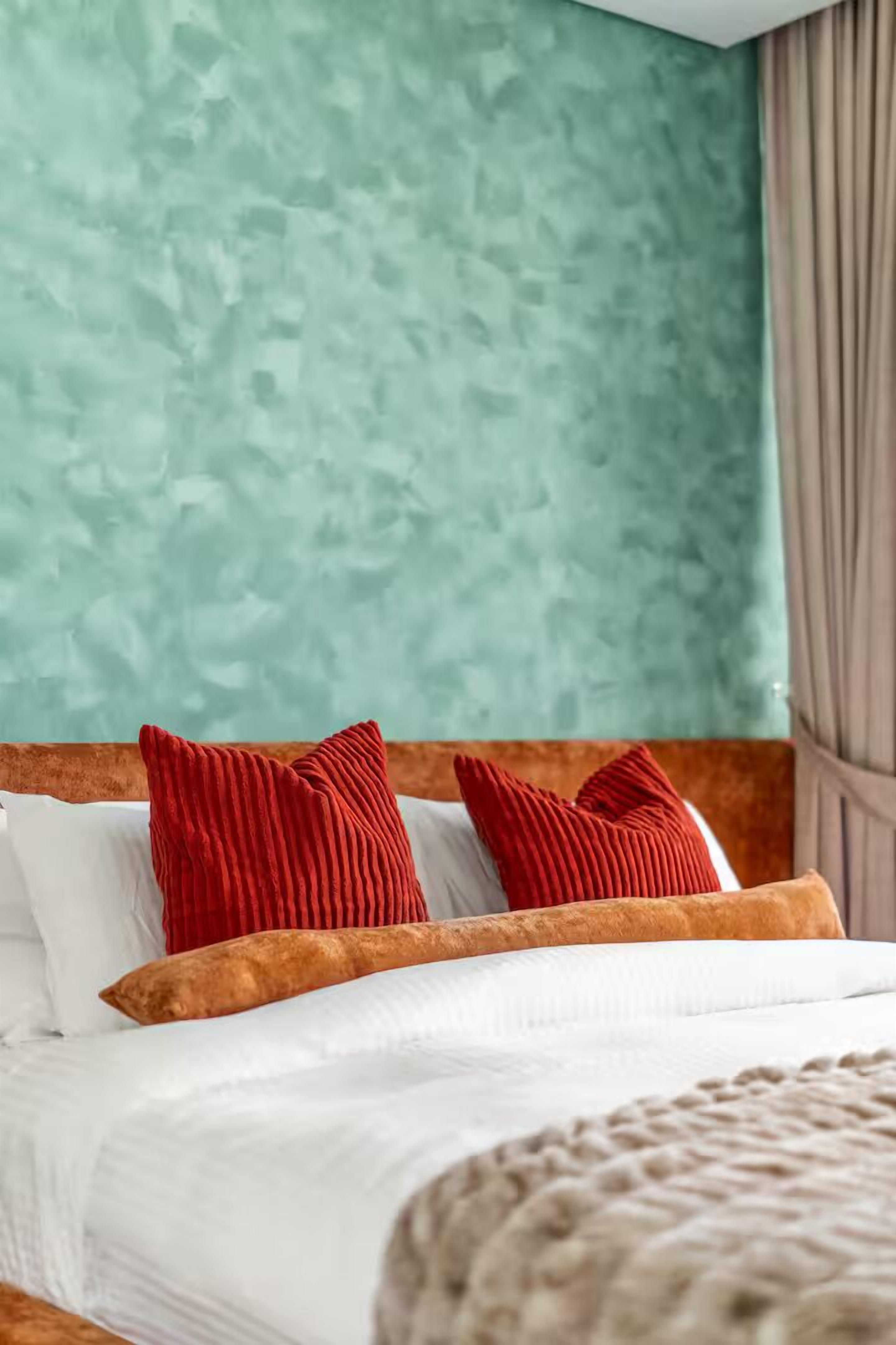 Una cama bien tendida con sábanas blancas y dos almohadas rojas, frente a una pared texturizada color turquesa.