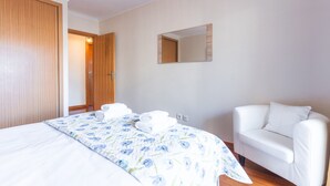 A bedroom with a bed, a white chair, a wooden wardrobe, and a mirror on the wall.